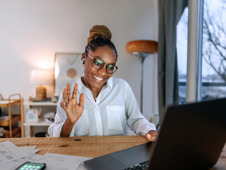A Black woman smiling and waving at her laptop.